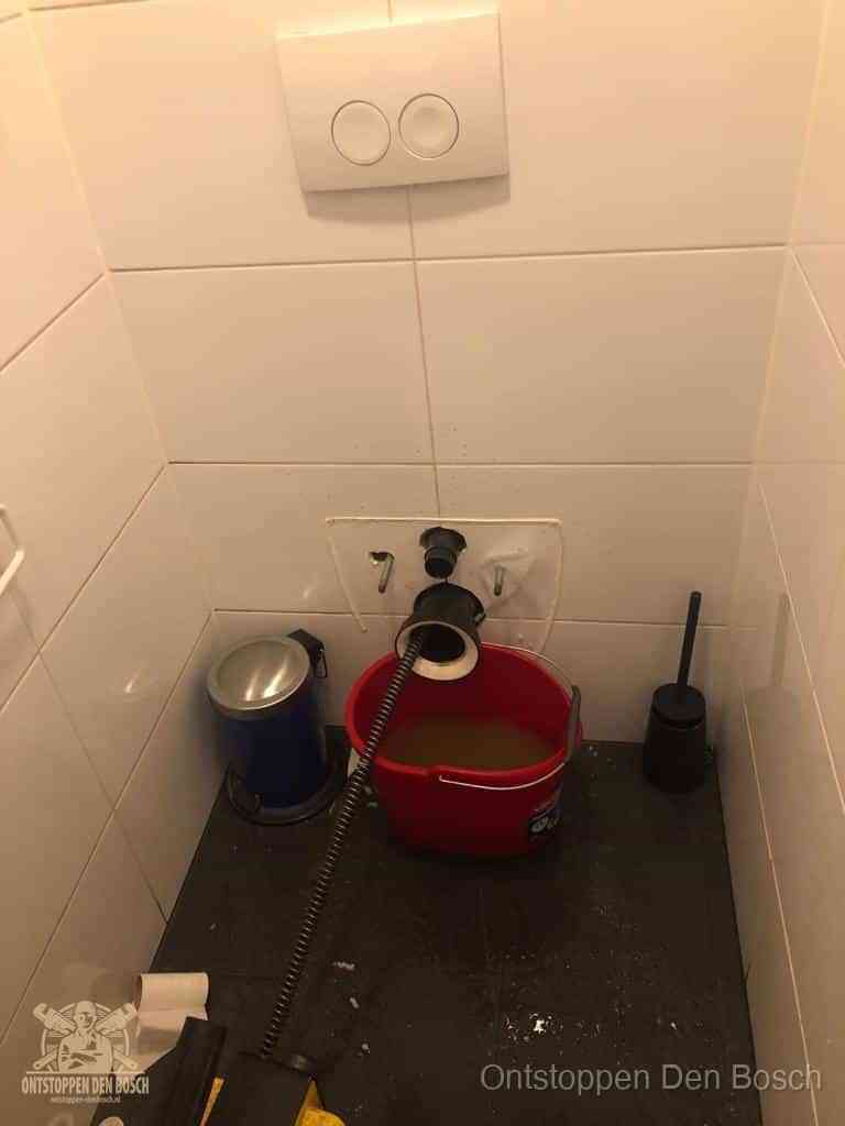 Een ontstopte wand-wc met verwijderde toiletpot waar een ontstoppingsveer in de wanduitgang verdwijnt, het achterhalen waar de verstopping zit in Den Bosch