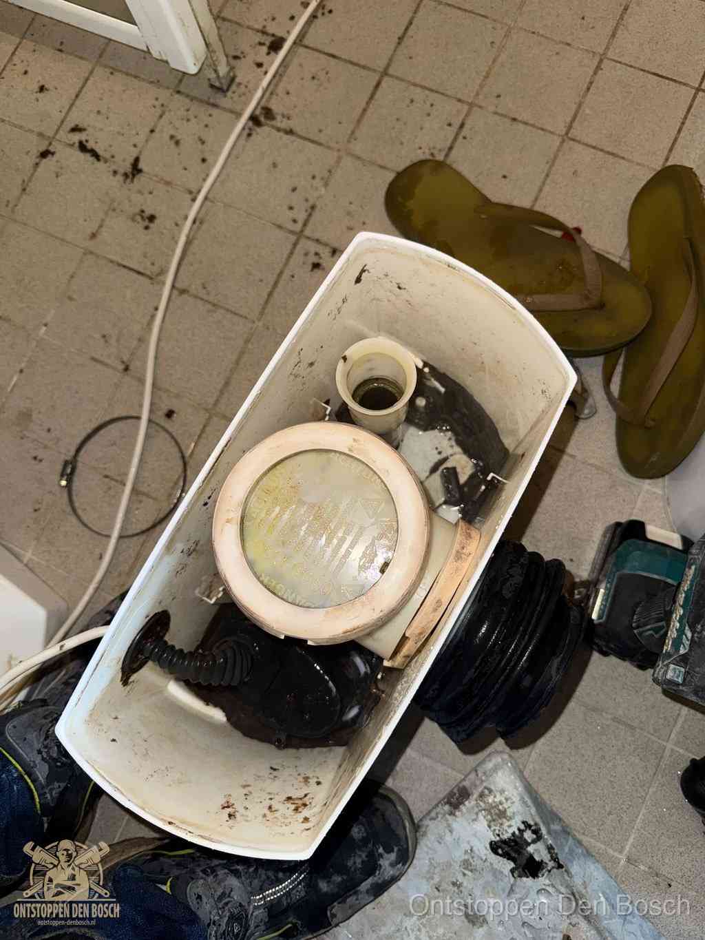 Een gedemonteerde toiletpot op de vloer met ontstoppingsveer en gereedschap ernaast, het verwijderen van een vastzittend voorwerp uit de wc in Den Bosch