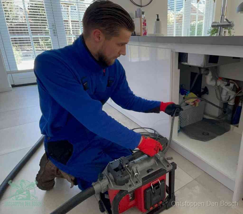 Een monteur met rode handschoenen knielt in een keuken en voert een elektrische ontstoppingsveer in tijdens het professioneel afvoer ontstoppen in Den Bosch
