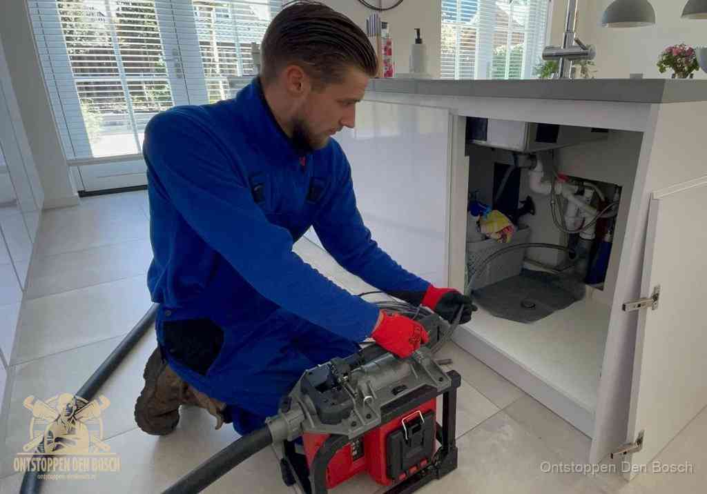 Een monteur voert een professionele elektrische ontstoppingsmachine in de afvoer onder de spoelbak tijdens het gootsteen ontstoppen in Den Bosch
